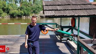 A man in a navy blue shirt is standing on a wooden dock near the water with a lifesaver hanging on the side.
