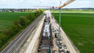 Luftaufnahme einer Baustelle für Eisenbahngleise mit Kränen, Maschinen und Arbeitern inmitten grüner Felder und städtischem Hintergrund.