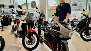 Ein Mann steht neben einem Motorrad in einer Ausstellung, in der mehrere Motorräder geparkt sind. Er lächelt und trägt ein blaues Hemd mit einem Logo auf der linken Brust.