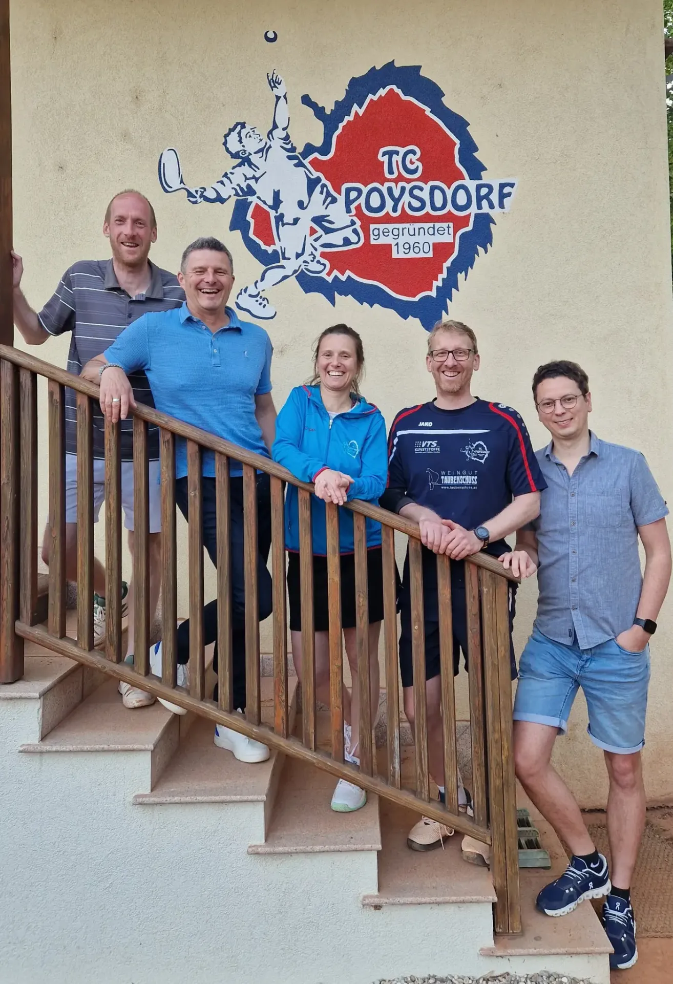 Fünf Menschen stehen auf einer Treppe. Drei Männer und zwei Frauen. Sie lächeln für ein Foto. Ein Logo ist an der Wand hinter ihnen.