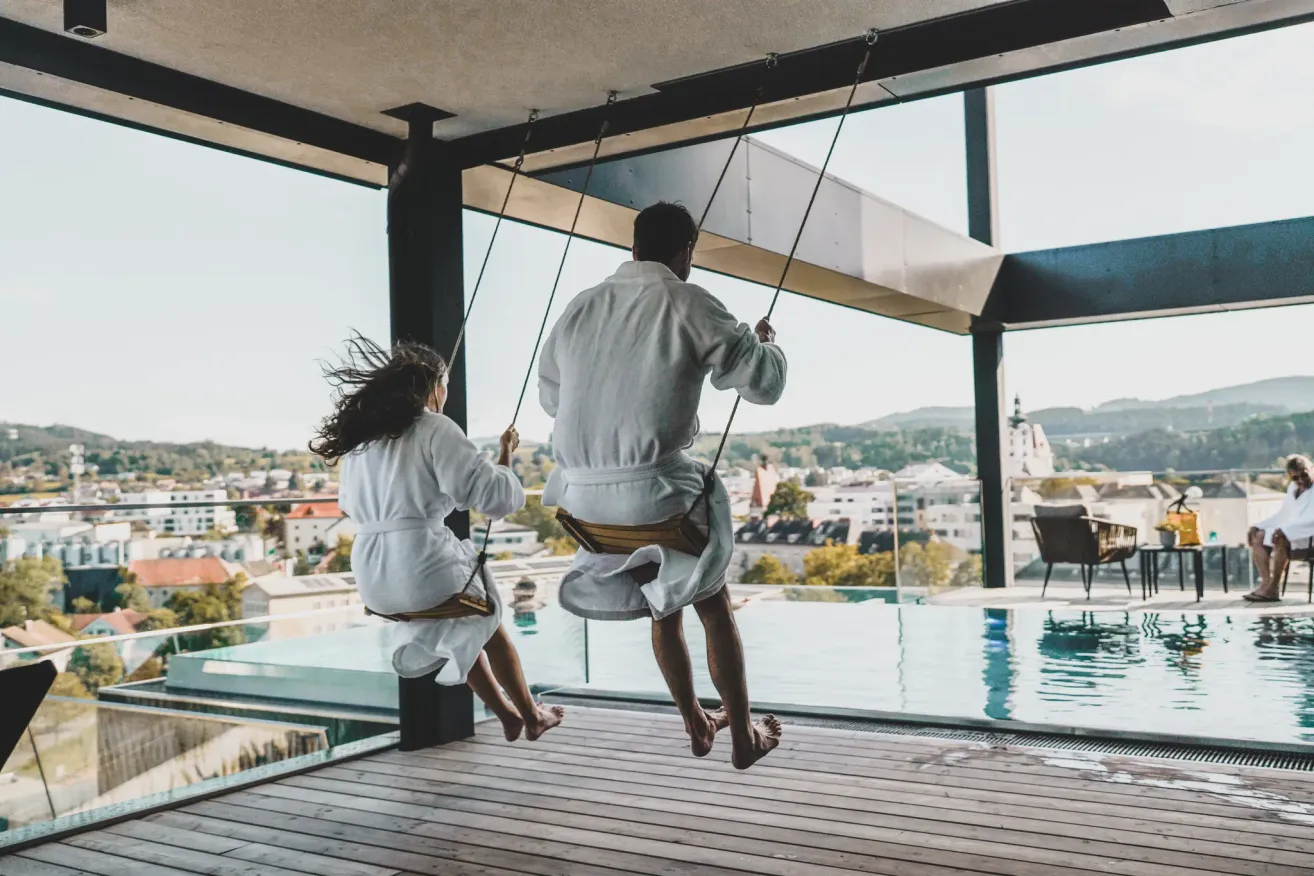 Two individuals in bathrobes swing on outdoor swings, overlooking a cityscape with buildings and mountains.