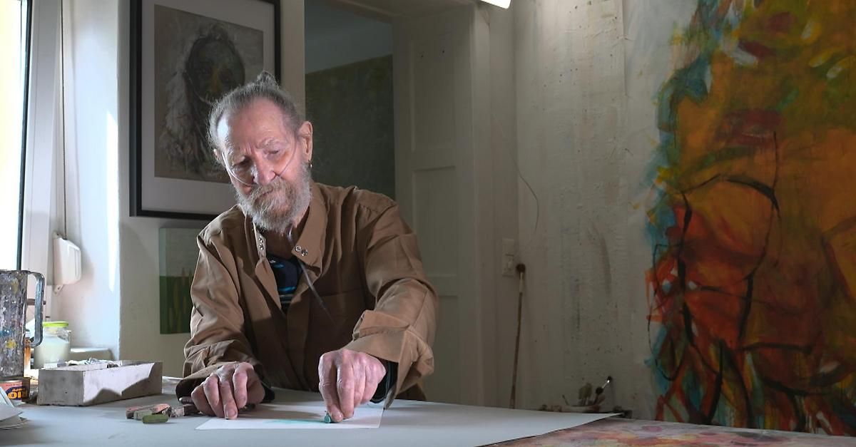 Ein älterer Mann mit Bart arbeitet in einem Studio an einem Blatt Papier. Der Raum hat abstrakte Gemälde an den Wänden.