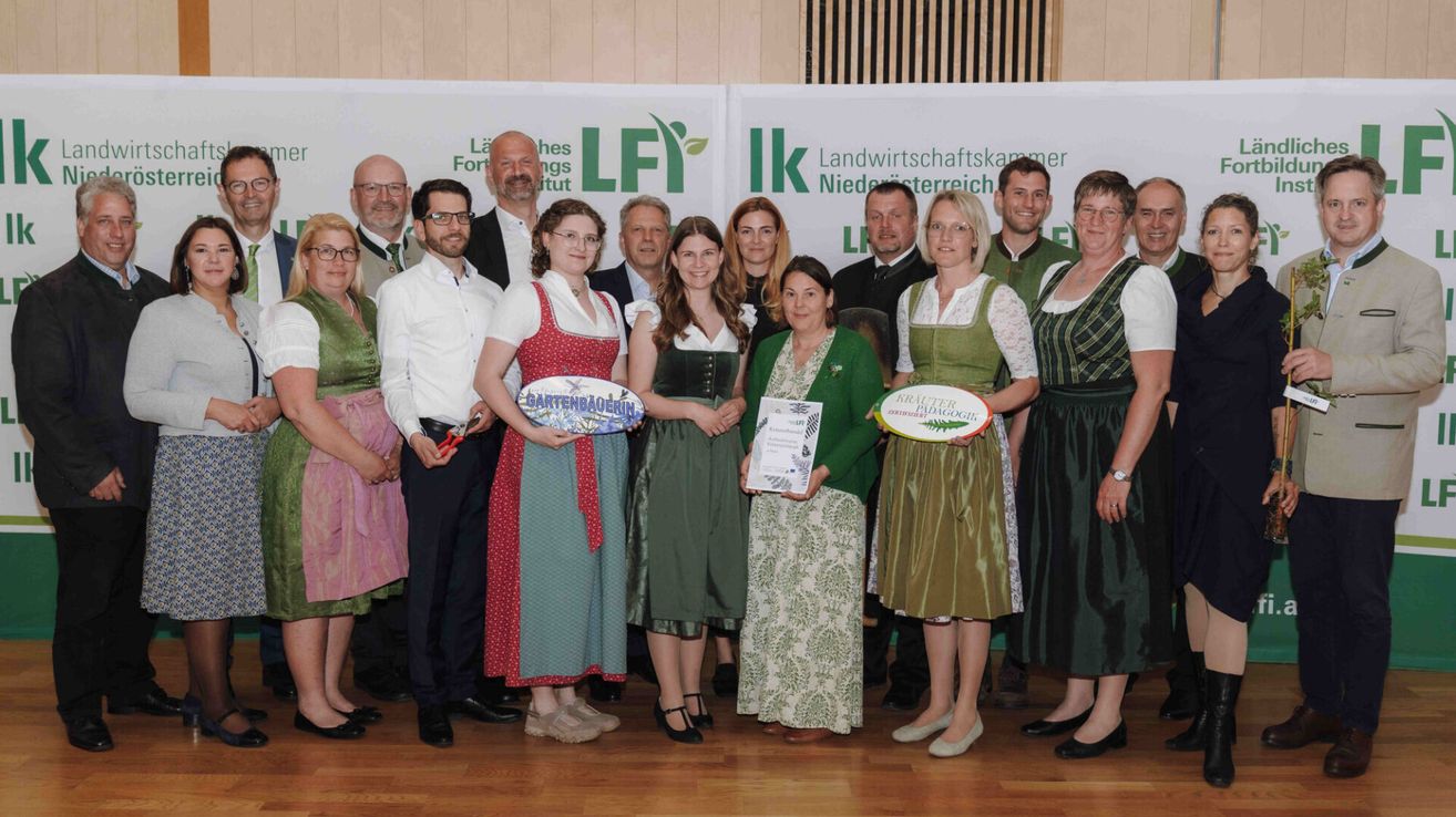 A group of people are posing for a picture while holding trophies. They are wearing traditional clothes. The woman in the center is holding a certificate. Behind them is a backdrop with logos and text.