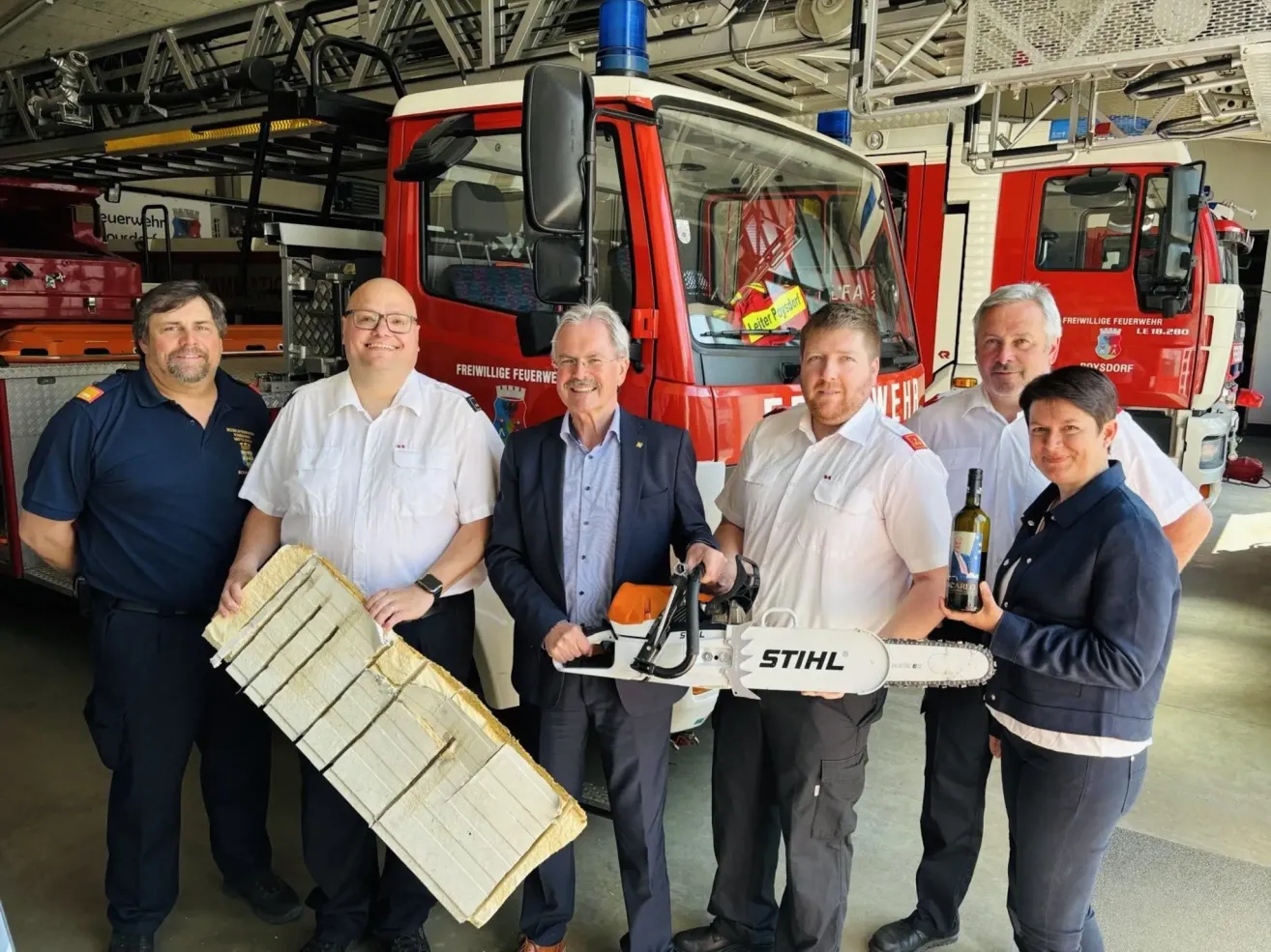 Eine Gruppe Feuerwehrleute steht vor einem Feuerwehrauto, hält ein großes Stück Holz und eine Kettensäge. Ein Mann in Anzug hält die Kettensäge, und eine Frau hält eine Flasche.