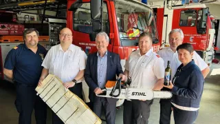 Eine Gruppe Feuerwehrleute steht vor einem Feuerwehrauto, hält ein großes Stück Holz und eine Kettensäge. Ein Mann in Anzug hält die Kettensäge, und eine Frau hält eine Flasche.