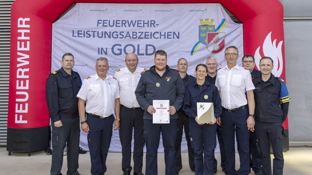 Eine Gruppe Feuerwehrleute in Uniform steht vor einem Banner. Ein Mann in der Mitte hält ein Zertifikat und eine Frau neben ihm ein Buch.