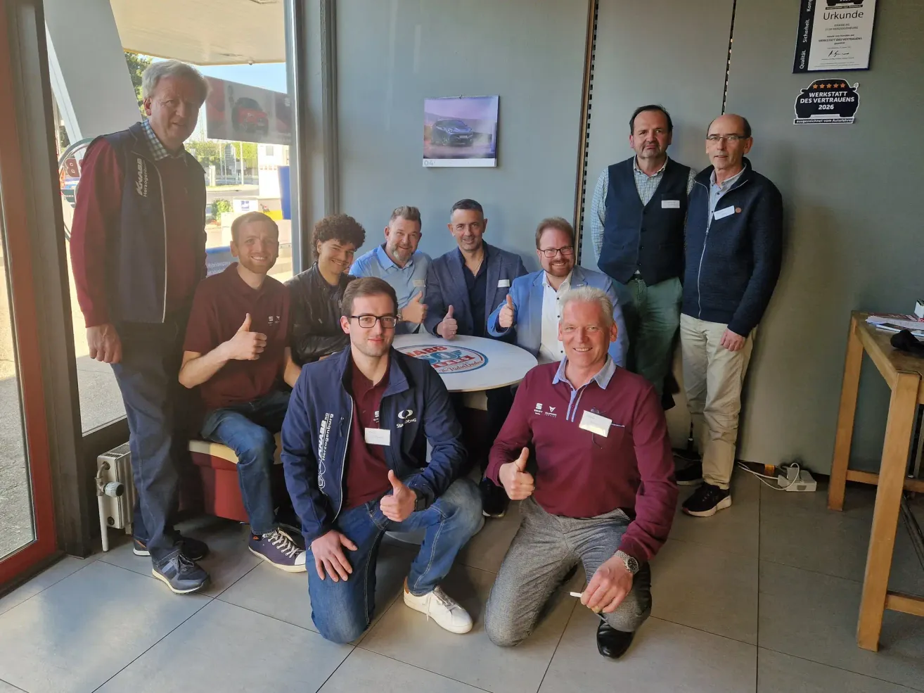 Eine Gruppe von Männern posiert für ein Foto in einer Ausstellung, einige kniend und andere stehend hinter einem Tisch mit einem Logo.