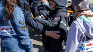 Ein erwachsener Mann in Uniform hilft einem Kind mit Polizeihelm, während ein weiteres Kind zuschaut. Sie sind draußen, mit einem Metallzaun im Hintergrund.