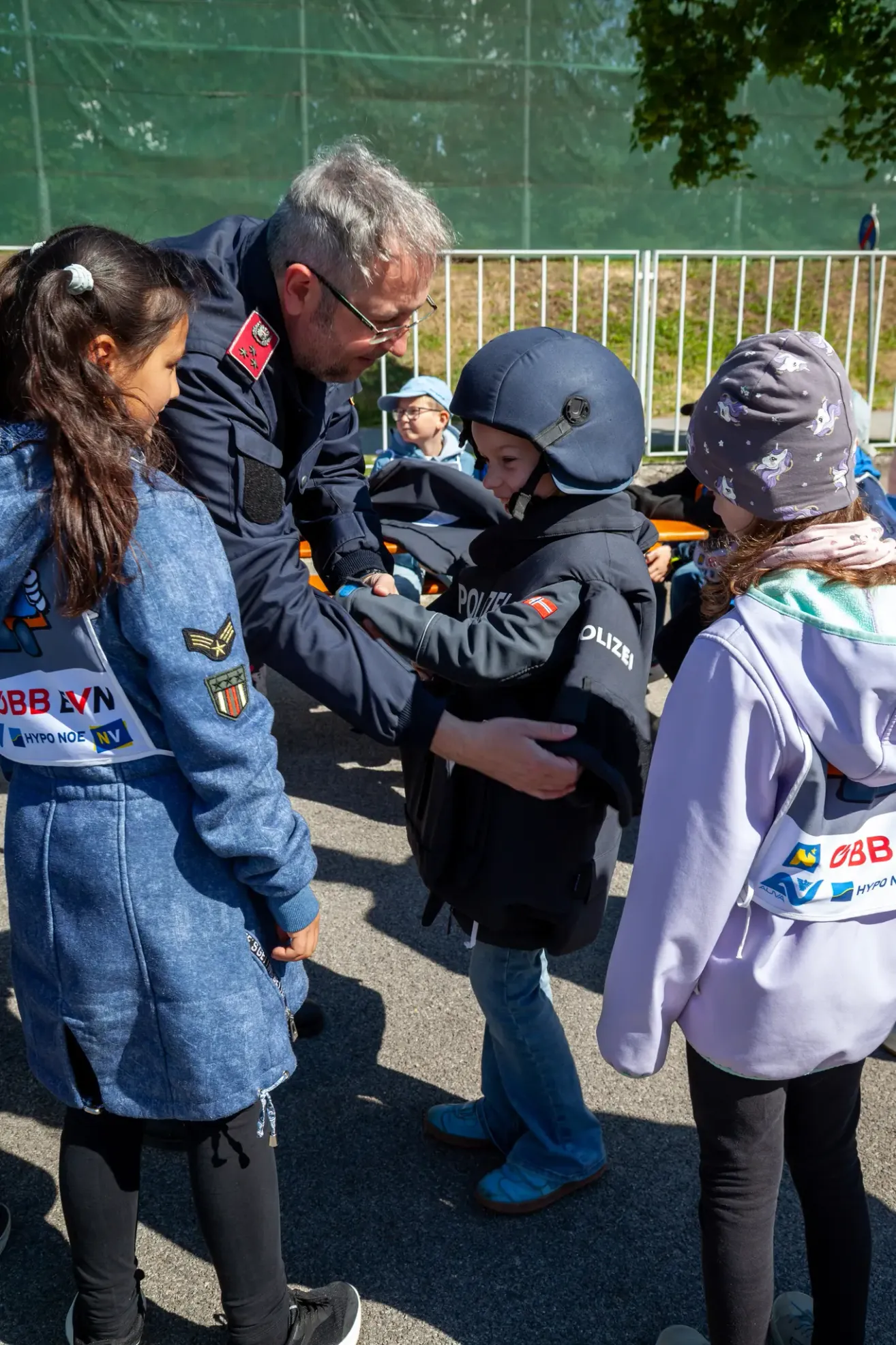 Ein erwachsener Mann in Uniform hilft einem Kind mit Polizeihelm, während ein weiteres Kind zuschaut. Sie sind draußen, mit einem Metallzaun im Hintergrund.
