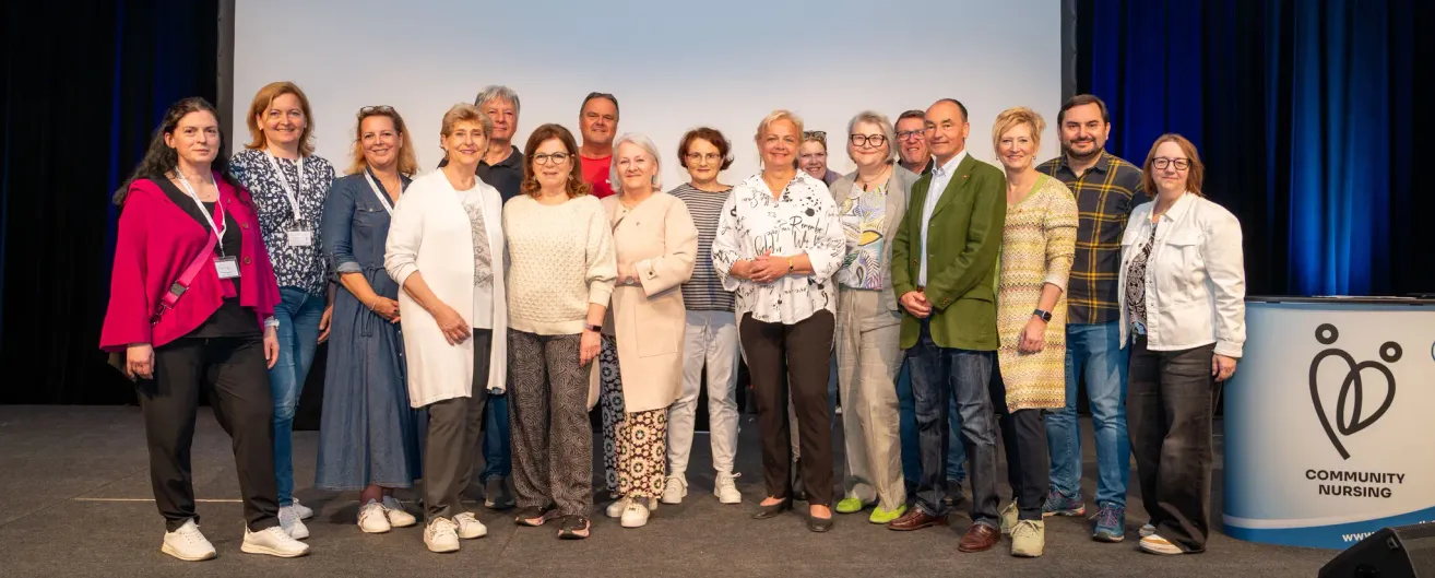Eine Gruppe von Menschen, möglicherweise ein Team, steht zusammen in einem Studio. Sie alle lächeln und scheinen für ein Foto zu posieren. Sie tragen formelle Kleidung.
