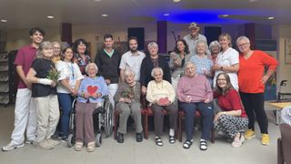 Eine Gruppe älterer Menschen, einige davon im Rollstuhl, posiert für ein Foto. Sie lächeln und halten herzförmige Karten. Einige Personen im Hintergrund stehen und lächeln. Eine Frau im Rollstuhl hält einen Blumenstrauß. Der Raum hat einen gefliesten Boden und blaue Lichter.