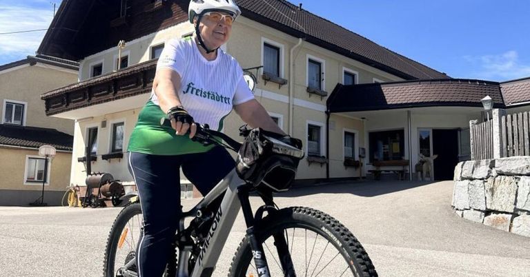A woman rides a mountain bike in front of a building. She wears a white helmet and a white shirt with the text Freistadter.