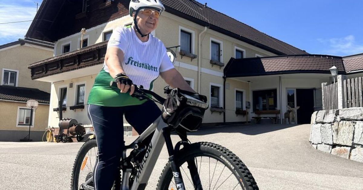 A woman rides a mountain bike in front of a building. She wears a white helmet and a white shirt with the text Freistadter.