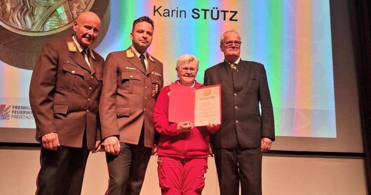 A woman in red holds a certificate on stage with three men in uniforms. One man wears glasses and a tie, another has a medal. A rainbow backdrop displays Karin Stutz.