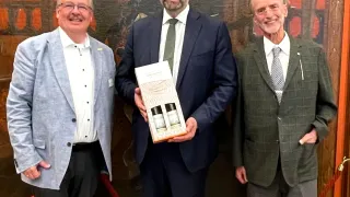 Drei Männer stehen nebeneinander und lächeln. Der Mann in der Mitte hält eine Box mit Parfüm. Hinter ihnen ist eine große Wandmalerei an einer Wand.