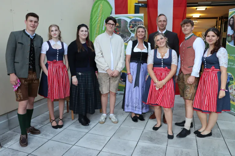 Eine Gruppe von Menschen in traditioneller deutscher Kleidung, die lächelnd für ein Foto vor einer Kulisse mit Bannern und Flaggen posieren.
