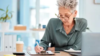 An elderly woman with glasses is sitting at a desk and writing on a piece of paper with a pen.
