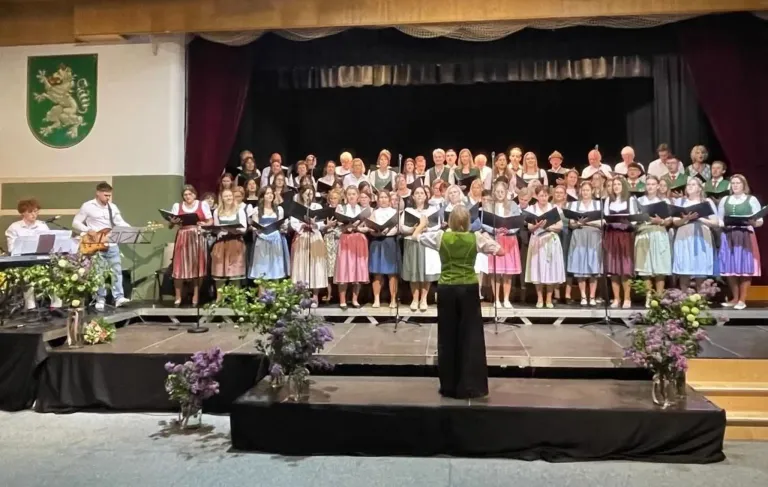 Ein Chor in traditioneller Kleidung tritt auf der Bühne auf, angeführt von einer Frau in Grün, mit Blumen am Bühnenboden.