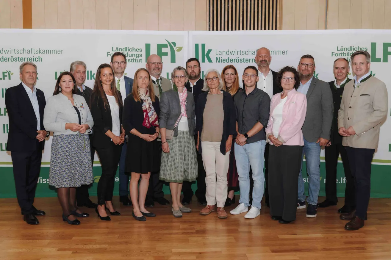A group of people stand together on a wooden floor, dressed in formal attire. Behind them, there are banners, one reading 'Landwirtschaftskammer Niederosterreich' and another featuring 'Ländliches Fortbildungsinstitut'.