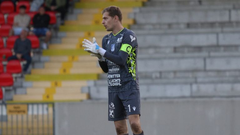 A soccer player wearing a jersey with the number 1 stands on a field. He wears gloves and has a focused expression. The background shows empty bleachers.