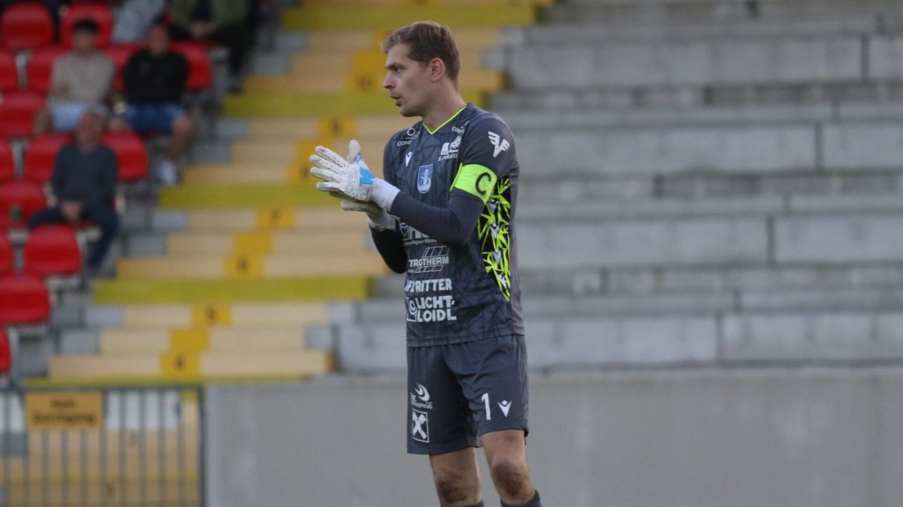 A soccer player wearing a jersey with the number 1 stands on a field. He wears gloves and has a focused expression. The background shows empty bleachers.