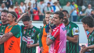 A group of soccer players in green and orange jerseys celebrate a victory. One holds a child, smiling with others. Spectators in the background watch eagerly.