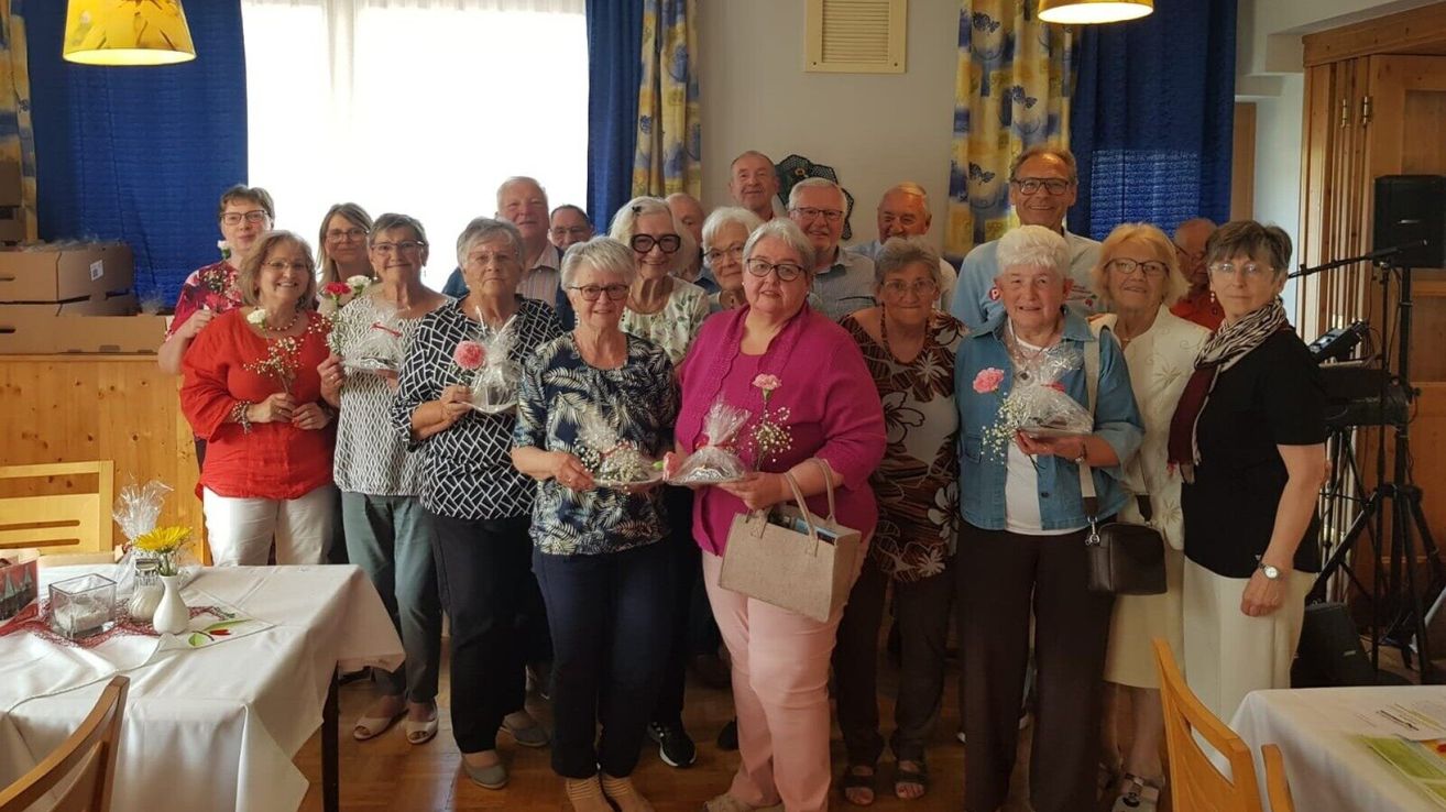 Eine Gruppe älterer Menschen steht zusammen und hält Geschenke. Sie tragen alle eine Brille. Die Frau im rosa Top hält einen Blumenstrauß.