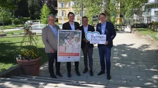 Vier Männer stehen auf einem Gehweg mit einem Plakat vor einem Gebäude und halten ein Schild, das Thermen & Vulkanland Steiermark zeigt. Sie sind schick gekleidet. Hinter ihnen befindet sich ein großes Gebäude mit gelben Wänden und einem Brunnen im Hintergrund.