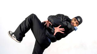 A man wearing a thick puffy black jacket is doing a martial arts move. He has one foot lifted off the ground.