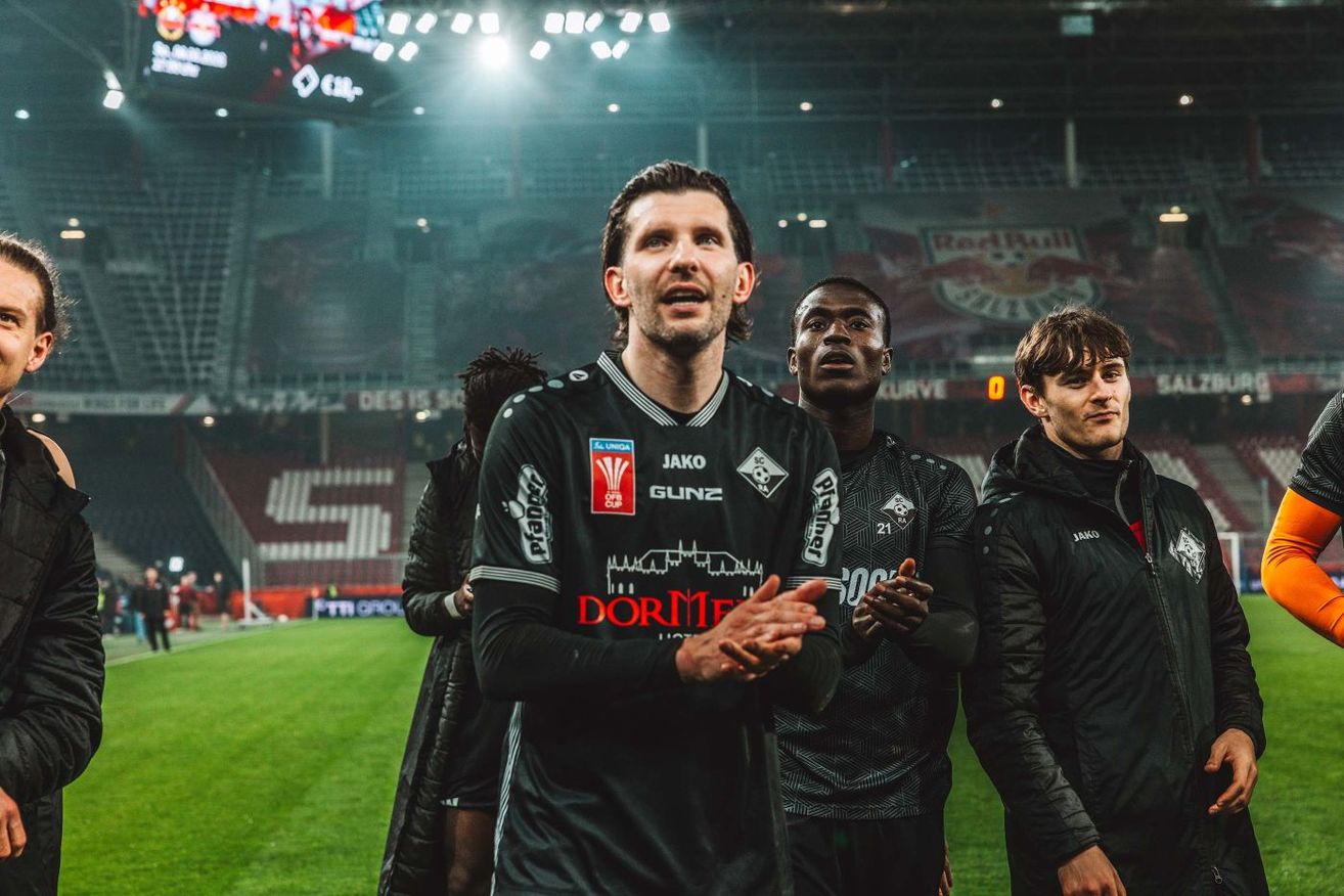 Four male athletes stand on a football field, likely after a match. One man, in the center, has his mouth open and hands clasped together. Behind him, three others look upwards. They wear black jerseys with white stripes. The background features an empty stadium with a redbull advertisement.