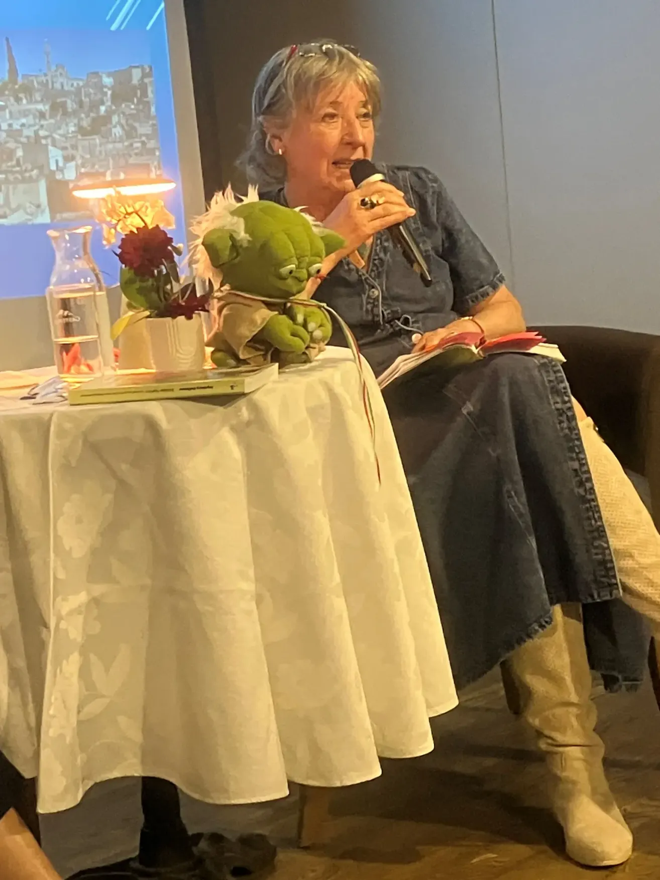 Eine Frau spricht in ein Mikrofon an einem Tisch. Sie trägt ein Jeanskleid und Stiefel. Ein Blumenvase und ein kleiner Plüschtier stehen auf dem Tisch. Ein Buch liegt auf ihrem Schoß.