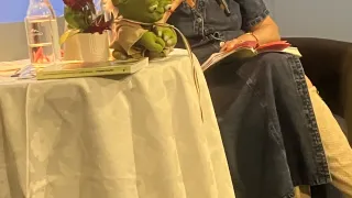 A woman speaks into a microphone at a table. She wears a denim dress and boots. A flower vase and a small plush toy are on the table. A book rests on her lap.
