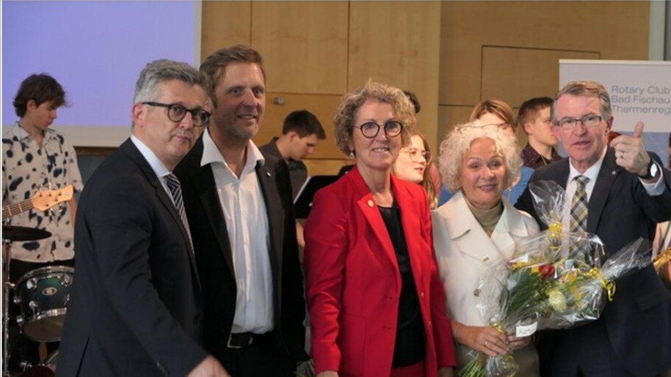 Five people, including three women, are smiling for a photo in a room. Two women are wearing glasses and holding flowers. The man on the left is wearing a tie.