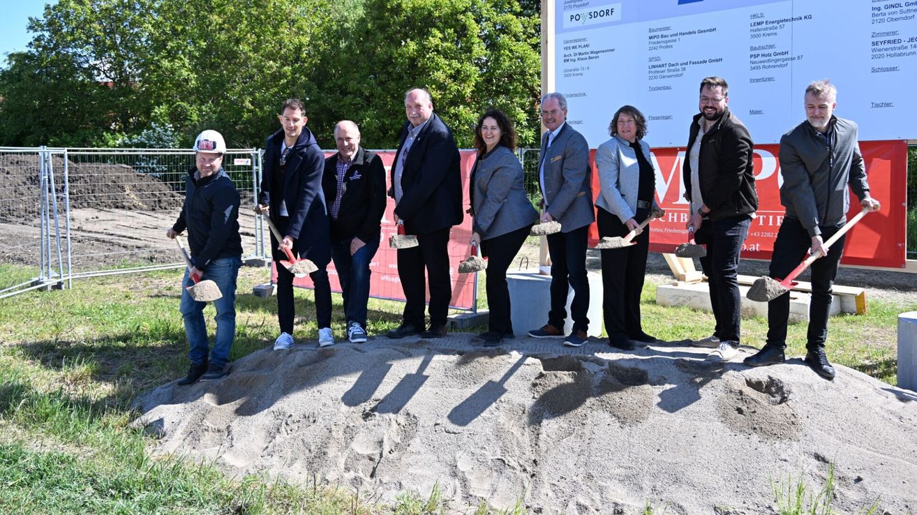 Mehrere Personen stehen an einer Baustelle und halten Schaufeln. Sie scheinen für ein Foto zu posieren. Ein Schild hinter ihnen sagt 'POSDORF'.