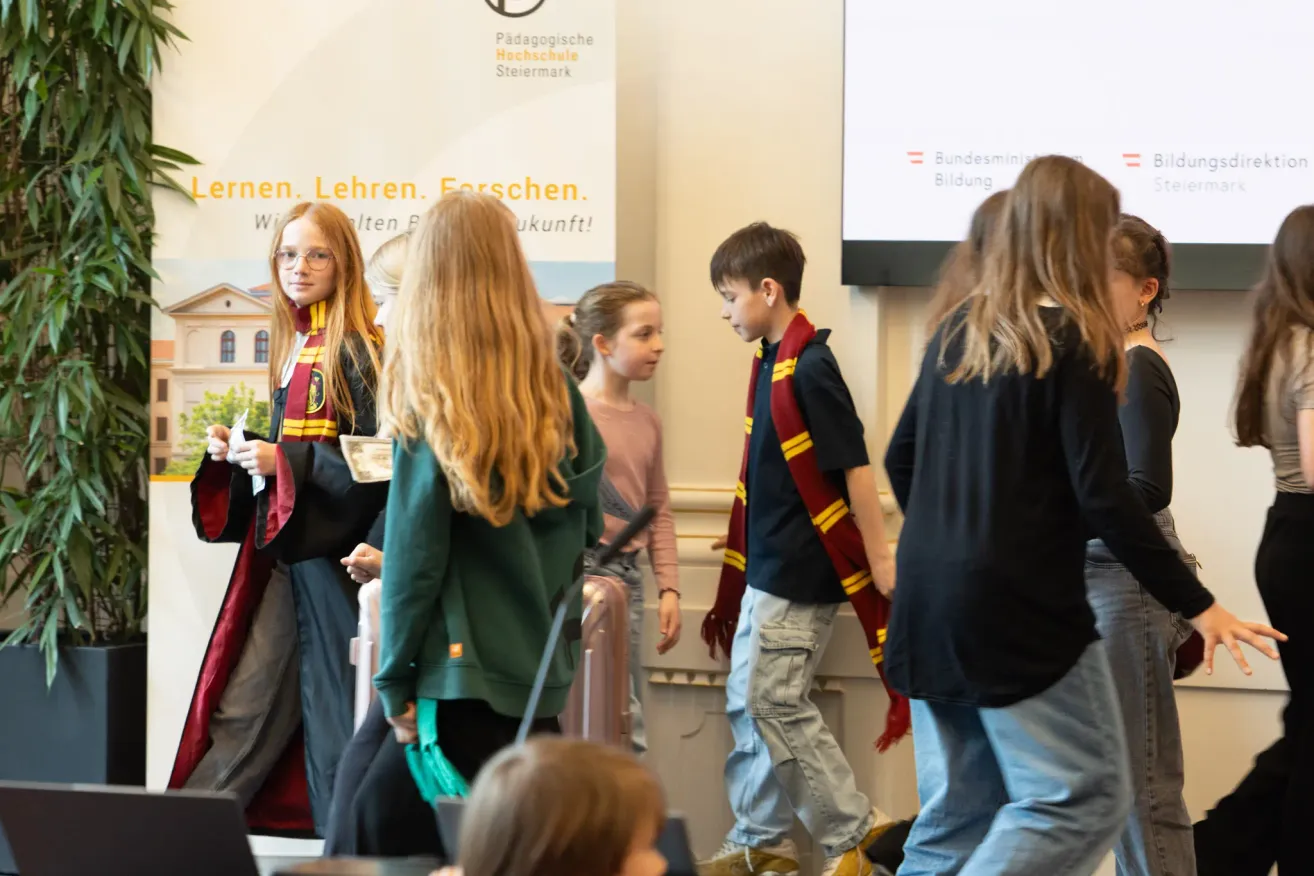 Mehrere Kinder in Schuluniformen stehen in einem Raum. Einige tragen Schals und eines hat eine Brille. Ein Banner mit dem Text 'Lernen. Forschen. Zukunft!' steht hinter ihnen.