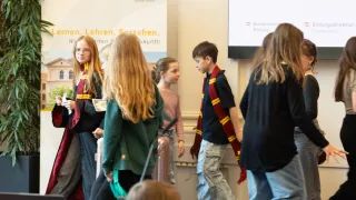 Several children in school uniforms are standing in a room. Some wear scarves, and one has glasses. A banner with the text 'Lernen. Forschen. Zukunft!' is behind them.