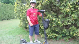 A young boy in a red shirt with the letter T stands next to an electric scooter with a black handlebar and bag, wearing a helmet and glasses.