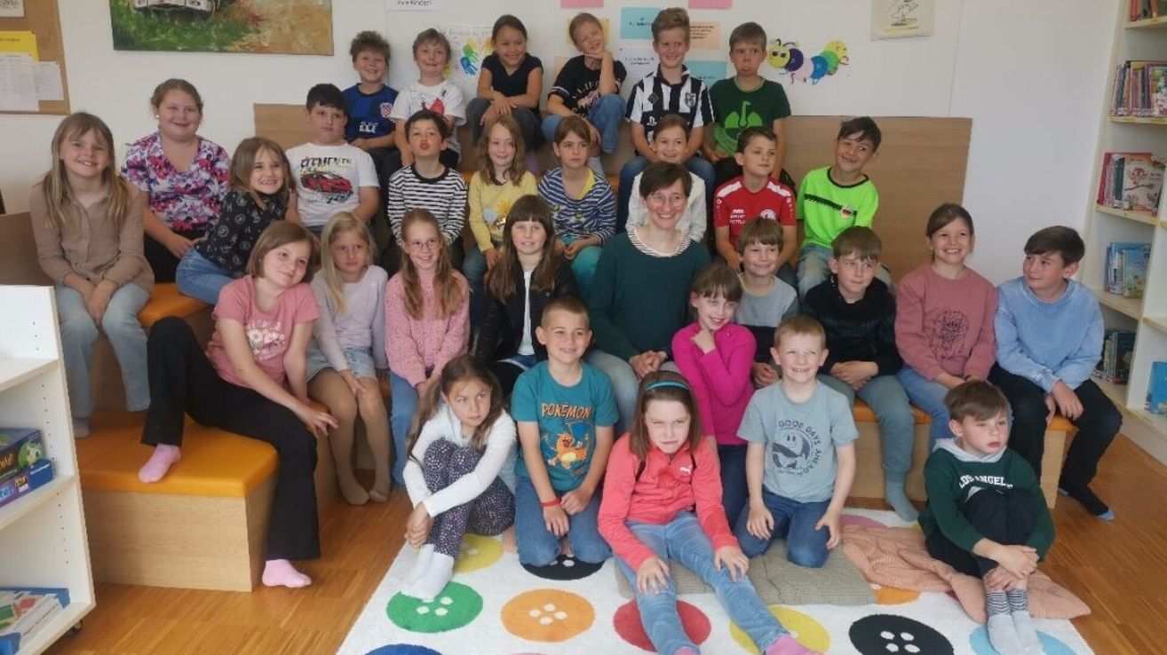 A group of children and an adult woman are sitting and standing on a bench and floor in a classroom. The children are wearing casual clothes. The woman is wearing glasses. The floor is covered with a colorful rug.