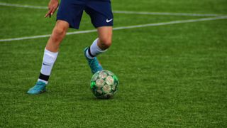 Ein Fußballspieler in blauen Shorts und blauen Nike-Schuhen tritt einen grün-braunen Fußball auf einem Rasen.