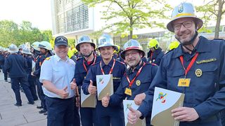Feuerwehrleute stehen draußen mit Helmen und Urkunden. Ein Mann gibt einen Daumen hoch. Ein Gebäude mit Glaswänden und einem Baum im Hintergrund.