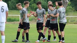 A group of young soccer players in gray jerseys are talking and interacting with each other on a grassy field.