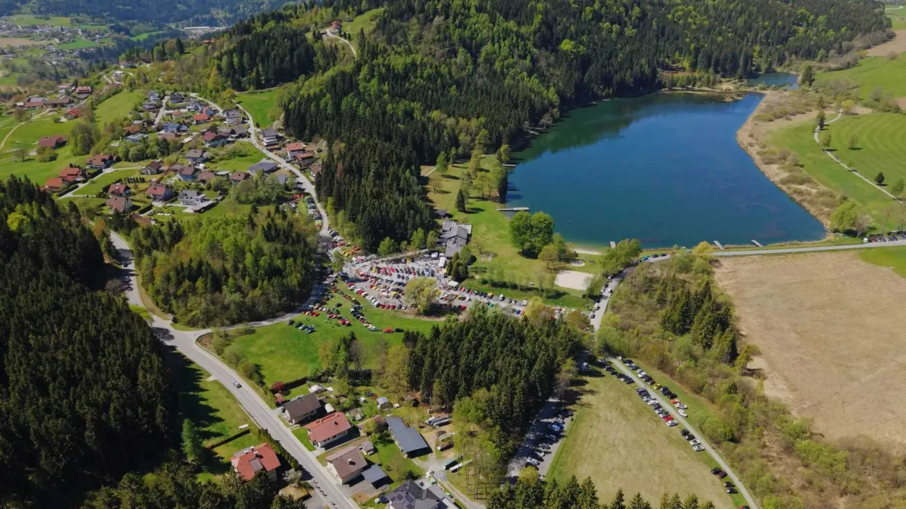 Luftaufnahme eines Sees, umgeben von Bäumen und Häusern, mit vielen geparkten Autos am Ufer.