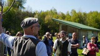 A man wearing a beret and glasses points to a tree as a group of people listen, standing outdoors near a wooden building.