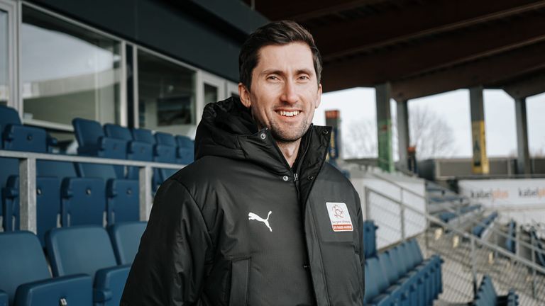 Ein lächelnder Mann in einer schwarzen Puma-Jacke posiert in einem Sportstadion mit blauen Sitzen und einem klaren Himmel im Hintergrund.