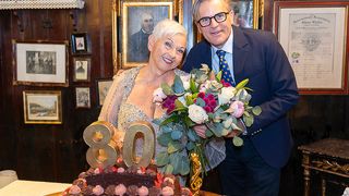 An elderly woman and a man stand by a cake with the number 80, holding a bouquet of flowers, with a golden trophy nearby. Behind them is a wall with framed pictures and a wooden shelf.