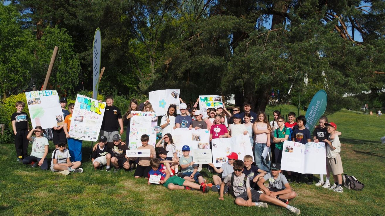 A group of children and adults are posing for a photo in a park with signs, trees in the background.