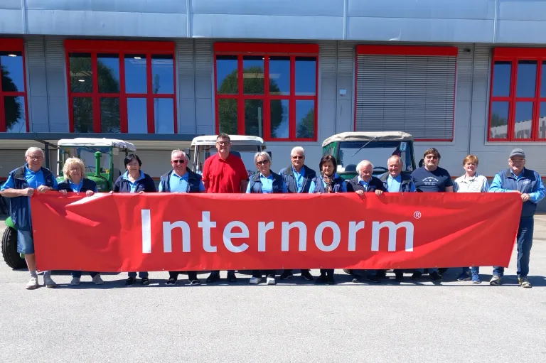 Eine Gruppe von Menschen steht hinter einem roten Banner mit dem Schriftzug Internorm vor einem Gebäude. Sie tragen blaue Hemden. Dahinter stehen zwei Fahrzeuge mit grünen Dächern.