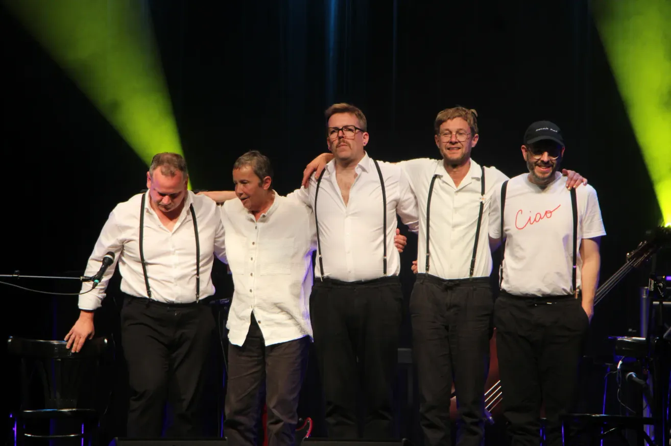 Five men in white shirts and black suspenders stand on stage. They are smiling and appear to be posing for a photo. Green lighting is in the background.