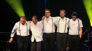 Fünf Männer in weißen Hemden und schwarzen Hosenträgern stehen auf der Bühne. Sie lächeln und scheinen für ein Foto zu posieren. Grüne Beleuchtung im Hintergrund.