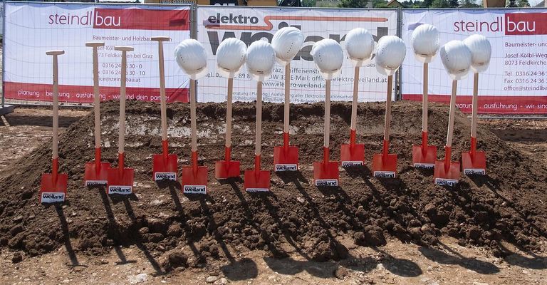 A line of red shovels with white helmets are placed in a row on the ground. Behind the shovels is a banner with the word "Elektro" and a company logo.